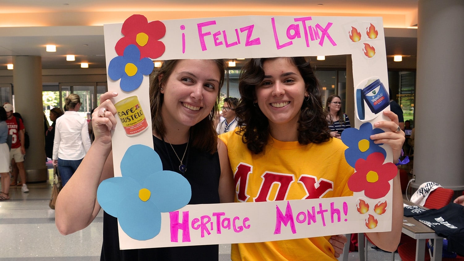 Two NC State students smile in the frame of a cardboard sign that reads "Feliz Latinx Heritage Month."